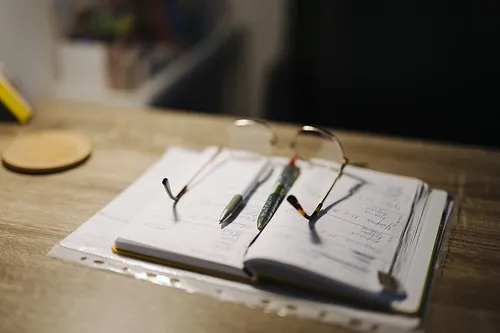 A person is studying at home during the evening. A notebook lies open on the desk with several pens. Reading glasses rest on the notebook, i